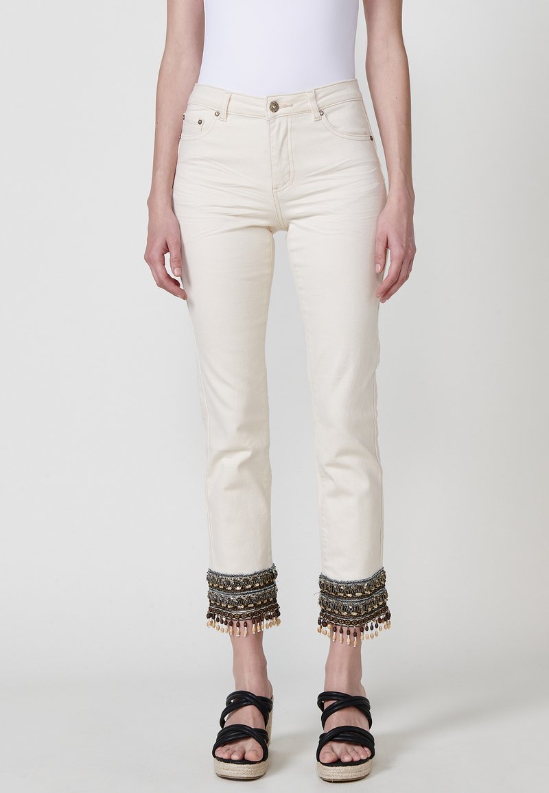Jeans de denim color crema con acentuaciones de cuentas y flecos en los puños, que presentan un diseño clásico de cinco bolsillos y cierre con cremallera.