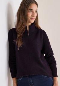 Lila gerippter Strickpullover mit hohem Ausschnitt und langen Ärmeln, kombiniert mit dunklen Jeans. Glatte Textur, lockere Passform.