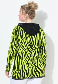 Langarm hoodie met een levendig groen en zwart zebra-streep patroon, gemaakt van lichte stof met een gestructureerde zwarte capuchon.