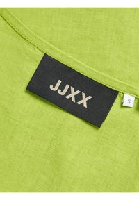 Jarko zelena tkanina s mekom teksturom; crna oznaka sadrži marku "JJXX" u svijetlo sivoj boji, uz oznaku veličine "S" sa strane.