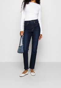 Jeans de tiro alto azul oscuro combinados con una blusa blanca de manga larga; los accesorios incluyen un pequeño bolso de mezclilla y bailarinas blancas.