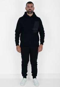 Felpa nera con cappuccio e tasca anteriore, caratterizzata da un tessuto testurizzato e un accento lucido. Abbinata a pantaloni jogger neri e sneakers bianche.