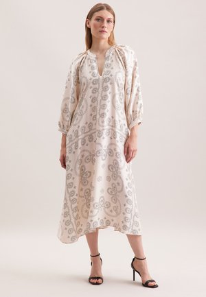 Langes, hellfarbenes Kleid mit Paisleymuster, V-Ausschnitt, Puffärmeln und fließendem Schnitt. Kombiniert mit schwarzen High-Heels.