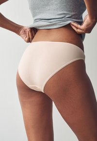 Šviesiai smėlio spalvos medvilninės trumpikės su lygiu paviršiumi ir subtiliu siuvimu, parodytos iš nugaros, atskleidžiančios vidutinio aukščio kirpimą ir prigludusį dizainą.