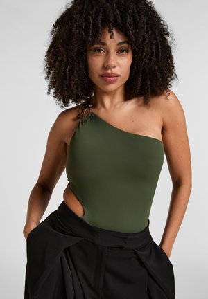 Femme aux cheveux bouclés portant un top asymétrique vert olive avec une découpe et un pantalon noir taille haute, debout les mains dans les poches.