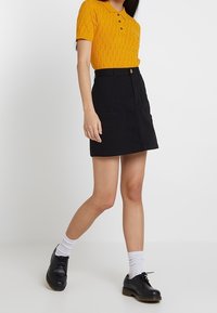 Polo shirt giallo a maglia con maniche corte e colletto, abbinato a una gonna di jeans nera con tasche frontali e scarpe nere con lacci indossate con calzini bianchi.