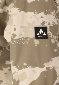 Waterdichte jas in beige met een witte spetterpatroon; voorzien van een zwart "Whistler" logo patch en een gladde, textuurstof.