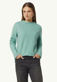 Helles türkisfarbenes Sweatshirt mit einer runden Ausschnitt und langen Ärmeln; kombiniert mit dunklen Jeans. Subtiles geometrisches Muster auf dem Stoff.