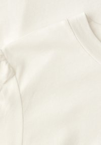 Close-up af blødt hvidt stof med en flæset skulderdetalje og ribstrikket rund hals på et beklædningsstykke.