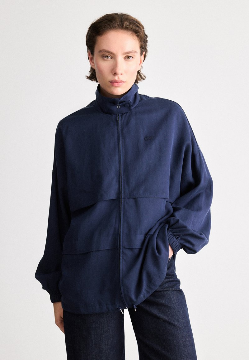 Giacca zippata blu navy con colletto alto, vestibilità ampia e maniche arricciate. Presenta una cucitura orizzontale e un sottile dettaglio del logo sul petto.