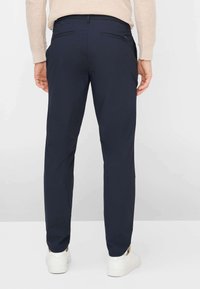 Pantaloni slim fit blu navy con finitura liscia, dotati di due tasche posteriori e indossati con sneakers bianche.