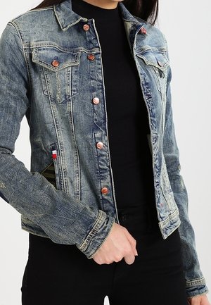 Chaqueta de mezclilla con botones de cobre, llevada sobre una camiseta de manga larga negra y pantalones negros, con la mano descansando cerca de la cintura.