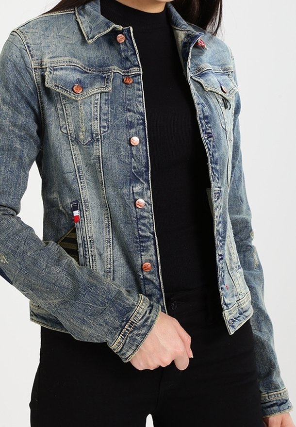 Jeansowa kurtka z miedzianymi guzikami noszona na czarnej bluzce z długim rękawem i czarnych spodniach, ręka spoczywająca w okolicy talii.