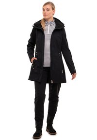 Veste imperméable noire avec capuche, poches zippées et coupe droite, associée à une chemise à rayures à manches longues et un pantalon sombre.