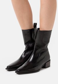 Botines de charol negro con puntera puntiaguda, tacón bajo y cierre de cremallera en la parte posterior. Textura suave y diseño minimalista.
