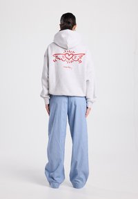 Sweat à capuche gris avec un graphisme de cœur ailé rouge au dos, associé à un pantalon ample bleu à rayures. Texture du tissu doux visible.