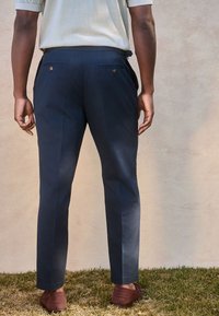 Pantalon bleu marine à coupe ajustée, doté de poches arrière avec boutons, associé à des mocassins marron. Tissu lisse et léger.