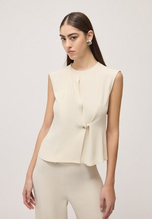 Giovane donna con lunghi capelli scuri indossa un top crema senza maniche con chiusura frontale e pantaloni coordinati su uno sfondo chiaro e semplice.