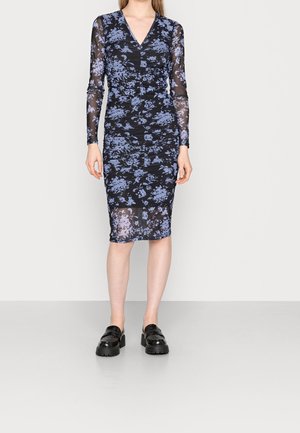 Mujer con un vestido negro ajustado con estampado floral azul y mangas largas transparentes, combinado con mocasines gruesos negros, de pie sobre un fondo liso.