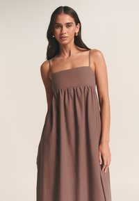 Bruine, vloeiende maxi-jurk met dunne bandjes, aansluitend lijfje en geplooide rok. Zachte textuur en effen kleur.