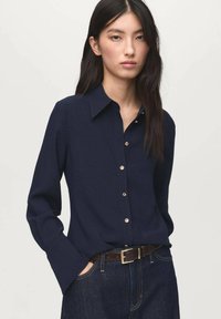 Navy long-sleeve shirt with a button-down collar and gold buttons, made from smooth fabric. Paired with high-waisted denim and a brown belt.