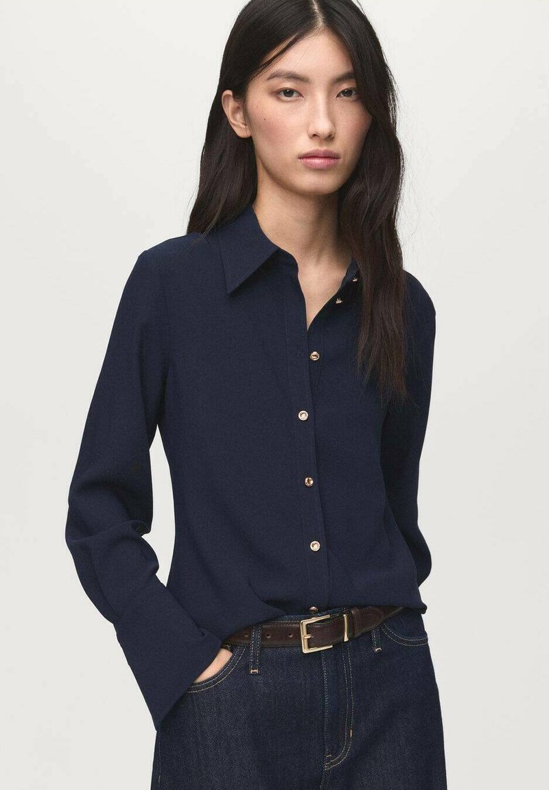 Navy long-sleeve shirt with a button-down collar and gold buttons, made from smooth fabric. Paired with high-waisted denim and a brown belt.