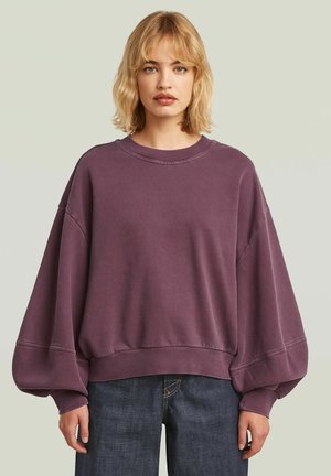 Donna con capelli biondi che indossa una felpa viola larga con maniche a palloncino e jeans blu scuro su uno sfondo chiaro uniforme.