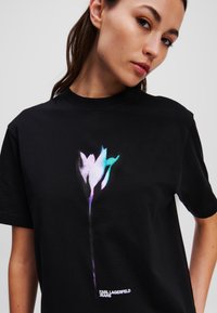 Juodas medvilninis marškinėliai su dideliu, stilizuotu gėlių grafiku mėlynos ir violetinės spalvų atspalviuose, o apačioje yra tekstas "KARL LAGERFELD JEANS".