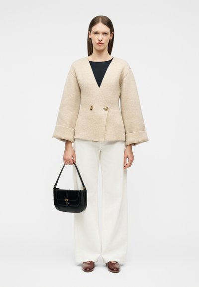 Γυναίκα με μπεζ σακάκι με διπλή σειρά κουμπιών, μαύρο τοπ, λευκό παντελόνι φαρδιάς γραμμής, καφέ παπούτσια, κρατώντας μαύρη τσάντα σε απλό φόντο.