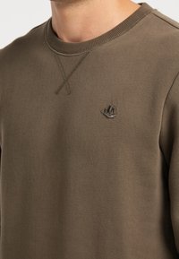 Dark olive green sweatshirt made of soft fabric, featuring a crew neck, cross-stitch detailing, and a small embroidered logo on the chest.