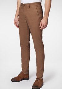 Brązowe bawełniane spodnie o slim fit, pięciopocketowym fasonie i szlufkach na pasek. W połączeniu z brązowymi sznurowanymi butami na neutralnym tle.