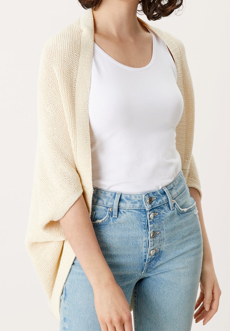 Femme portant un haut blanc sans manches, un cardigan écru en tricot, et un jean bleu clair taille haute avec quatre boutons visibles.