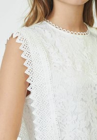 Top bianco in pizzo con intricate fantasie floreali, bordi scallop e delicati dettagli intrecciati lungo le maniche e il colletto.