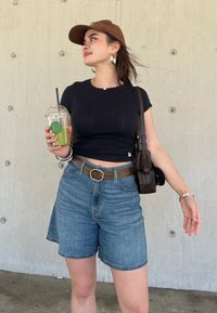 Svart åtsittande t-shirt, högmidjade denimshorts, brun bälte, brun keps, håller en genomskinlig kopp med grön dryck, läder crossbody-väska.