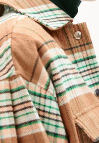 Närbild av ett tan-rutat tyg med gröna, vita och svarta ränder, som har en metalltryckknapp på vikta plaggskikt.