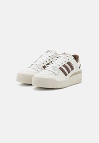 White sneakers with brown stripes, leather material, round toe, perforated toe box, and a thick, textured sole featuring a tread pattern.