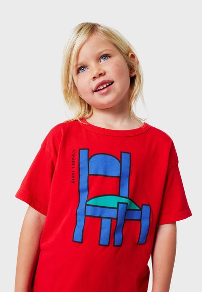 Rød bomulds T-shirt med et stort grafisk motiv af en stiliseret blå stol med en grøn sæde. Korte ærmer og rund hals.