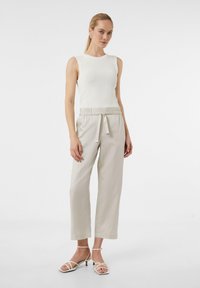 Biała ribbed bezrękawnik zestawiony z jasnobeżowymi spodniami na elastycznej talii. Modelka nosi minimalistyczne białe sandały na paski. Prosty, czysty design.