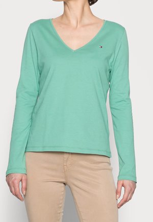 Frau trägt ein langärmliges, mintgrünes V-Ausschnittshirt mit kleinem Logo auf der Brust und beigen Hosen, steht vor einem einfarbigen Hintergrund.