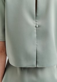 Blusa a maniche corte in morbido tessuto verde chiaro opaco con dettaglio a bottoni sul retro e design leggermente cropped. Texture liscia.