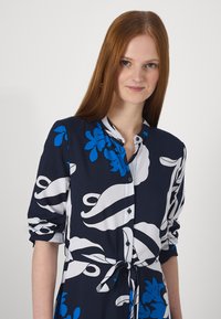 Vestido de marinha com padrão floral branco e azul, com botões à frente e mangas enroladas. Inclui um laço na cintura para ajuste. Tecido com textura suave.