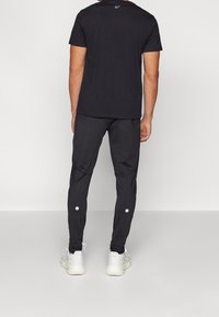 T-shirt noir à manches courtes avec un logo réfléchissant dans le dos, assorti à un pantalon noir avec des points réfléchissants près des chevilles, et des baskets claires.