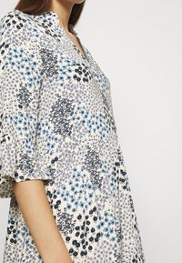 Kukkamekko, jossa on sekoitus sinisiä ja mustia kuvioita vaalealla taustalla, kauluksellinen pääntie ja röyhelödetaljeilla varustetut hihat.