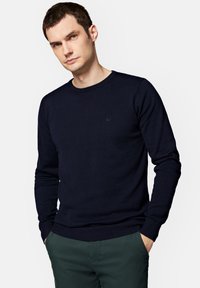 Granatowy sweter z dzianiny z okrągłym dekoltem, długimi rękawami oraz prążkowanymi mankietami i dołem. Posiada subtelne logo na klatce piersiowej.