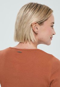 Femme blonde aux cheveux courts portant un haut côtelé couleur rouille, profil latéral visible, mettant en valeur des boucles d'oreilles à l'oreille gauche et un petit détail de collier.