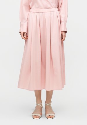 Nő rózsaszín, pliszírozott midi szoknyában, amelyhez ugyanilyen rózsaszín hosszú ujjú ing és fehér pántos szandálok társulnak.