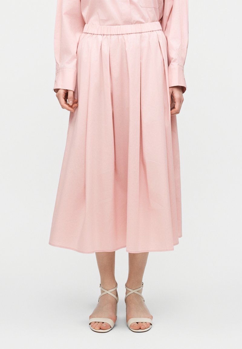 Γυναίκα που φοράει μίντι ροζ φούστα με πιέτες, συνδυασμένη με ροζ μακρυμάνικο πουκάμισο και λευκά σανδάλια με λουράκια.