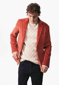 Oranje corduroy blazer over een crèmekleurige gebreide trui met een ruitpatroon, gecombineerd met donkere jeans. De stof vertoont zichtbare, gestructureerde lijnen.