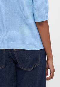 Suéter de malha azul claro com mangas curtas, apresentando uma textura canelada e uma bainha arredondada, combinado com jeans de ganga escura.