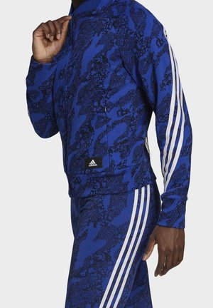 Homme portant un survêtement Adidas bleu et noir à motifs avec des bandes blanches sur les manches et les jambes du pantalon, ajustant le col de la veste.